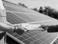 Przekazanie dokumentacji instalacji fotowoltaicznej bez zgłoszenia. Dwie ręce w rękawicach trzymają papiery na tle paneli słonecznych.