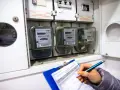 Kontrola liczników energii elektrycznej. Ręka z długopisem zapisuje dane na kartce, obok widoczne liczniki i bezpieczniki.