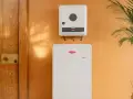 System magazynowania energii Fronius Symo GEN24 i akumulator Reserva na pomarańczowej ścianie, obok drewnianych drzwi i rośliny.