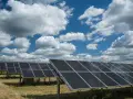 Rzędy paneli fotowoltaicznych 50kW na tle błękitnego nieba z białymi chmurami. W oddali widać turbiny wiatrowe.