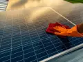 Ręka w pomarańczowej rękawiczce czyści panele fotowoltaiczne czerwoną ściereczką. Czyste panele to lepsza energia.