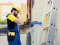 Certyfikowany instalator pomp ciepła w żółtym kasku i niebieskich ogrodniczkach przygotowuje się do pracy przy jednostce zewnętrznej.