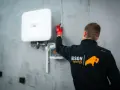 Technik w czerwonych rękawicach mierzy odległość od białego falownika. Działania te mają na celu zapobieganie kradzieży prądu.