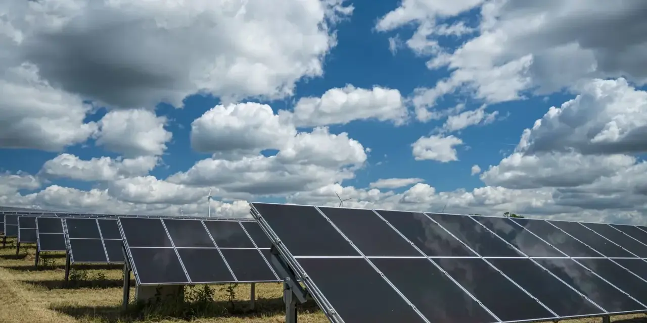 Rzędy paneli fotowoltaicznych 50kW na tle błękitnego nieba z białymi chmurami. W oddali widać turbiny wiatrowe.