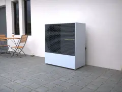 Nowoczesna pompa ciepła typu powietrze woda stoi na tarasie obok stolika z krzesłami.