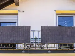 Dwa panele fotowoltaiczne na balkonie, obok okien z roletami i kwitnących kwiatów.