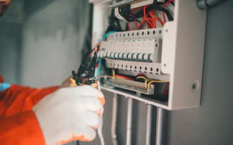 Elektryk w białych rękawicach przycina przewód do skrzynki z bezpiecznikami. Nie chce, by wybija bezpiecznik bez obciążenia.