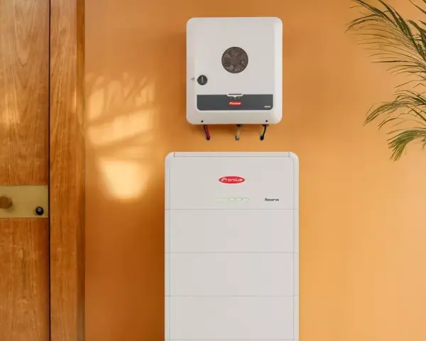 System magazynowania energii Fronius Symo GEN24 i akumulator Reserva na pomarańczowej ścianie, obok drewnianych drzwi i rośliny.