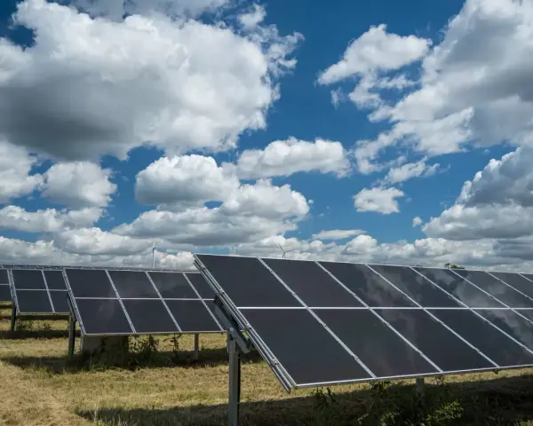Rzędy paneli fotowoltaicznych 50kW na tle błękitnego nieba z białymi chmurami. W oddali widać turbiny wiatrowe.