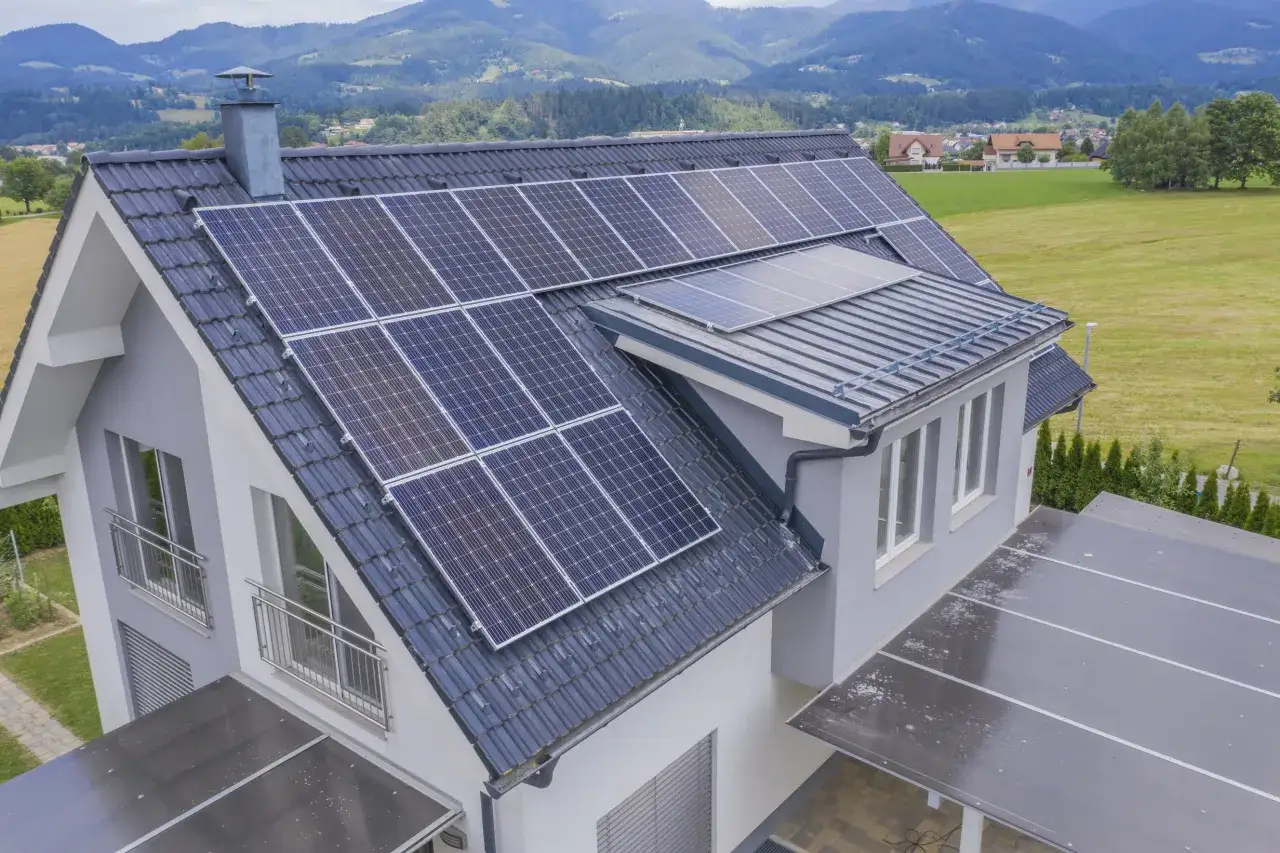 Nowoczesny dom z panelami fotowoltaicznymi na dachu, zapewniającymi bilansowanie energii. W tle góry i zielone pola.
